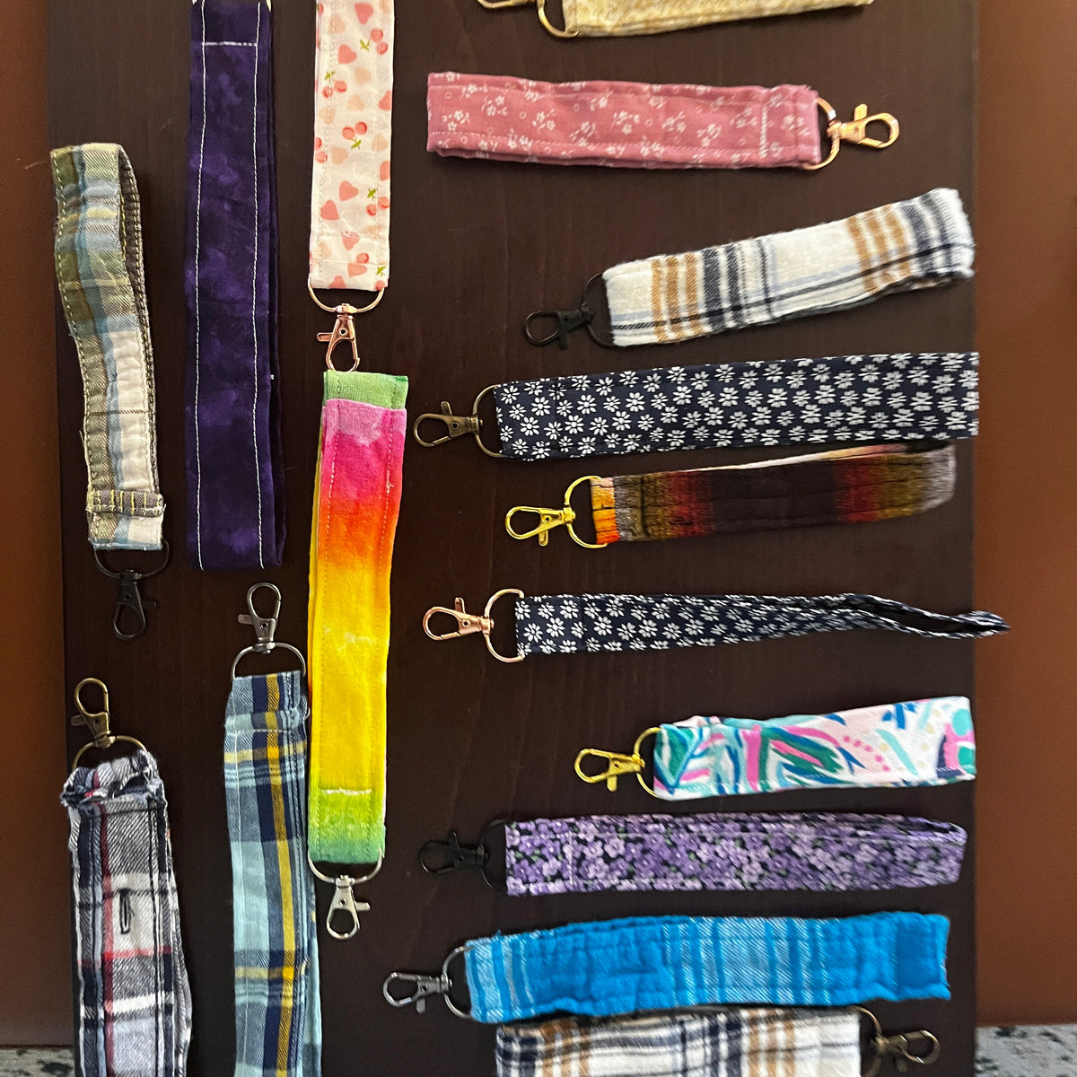 A collection of colorful fabric wristlets with metal lobster clasps, arranged vertically and horizontally on a dark wooden surface.